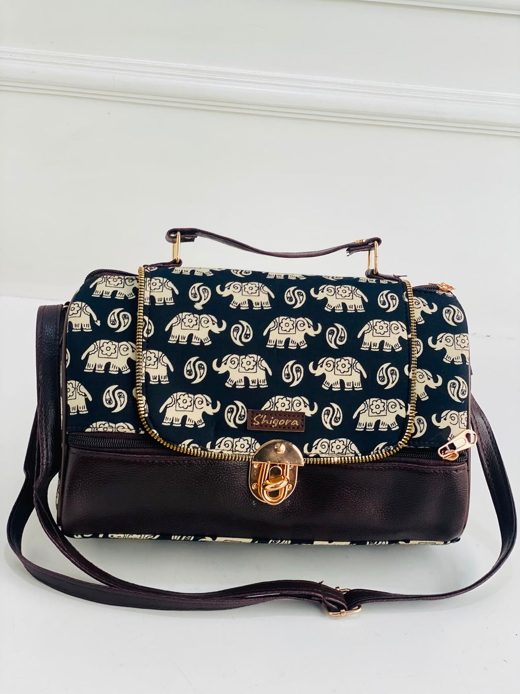 A navy blue handbag with a repeating cream elephant print on the flap and body, finished with dark brown leather trim, a top handle, and a gold-tone turn-lock clasp. The brand name 'Shagawa' is visible on a small leather patch.