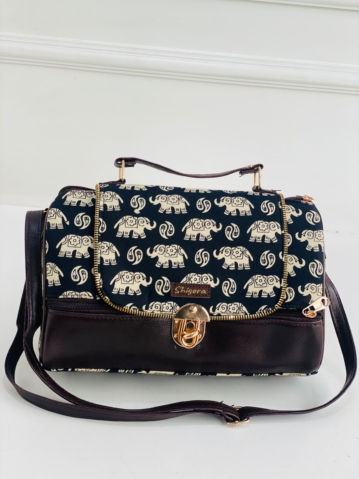 A navy blue handbag with a repeating cream elephant print on the flap and body, finished with dark brown leather trim, a top handle, and a gold-tone turn-lock clasp. The brand name 'Shagawa' is visible on a small leather patch.