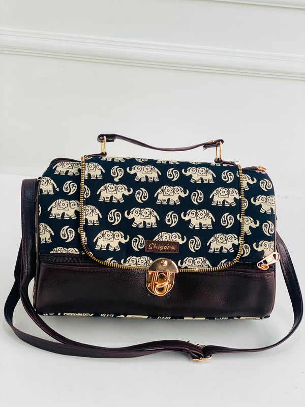 A navy blue handbag with a repeating cream elephant print on the flap and body, finished with dark brown leather trim, a top handle, and a gold-tone turn-lock clasp. The brand name 'Shagawa' is visible on a small leather patch.