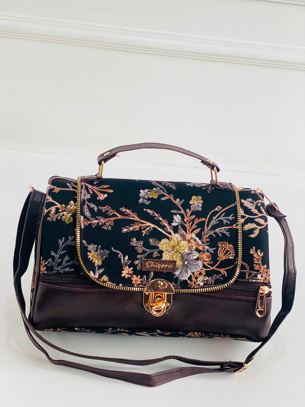 A dark brown leather handbag with a black floral embroidered flap, featuring gold-tone hardware, a top handle, and a detachable shoulder strap, set against a white paneled background.