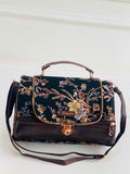 A dark brown leather handbag with a black floral embroidered flap, featuring gold-tone hardware, a top handle, and a detachable shoulder strap, set against a white paneled background.