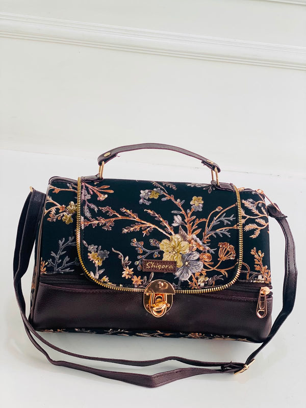 A dark brown leather handbag with a black floral embroidered flap, featuring gold-tone hardware, a top handle, and a detachable shoulder strap, set against a white paneled background.