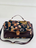 A structured handbag with a dark floral patterned flap and base, combined with a solid dark brown leather lower section and handle. It features gold-tone hardware, including a central turn-lock clasp and zipper pulls, and is displayed on a clean white background.