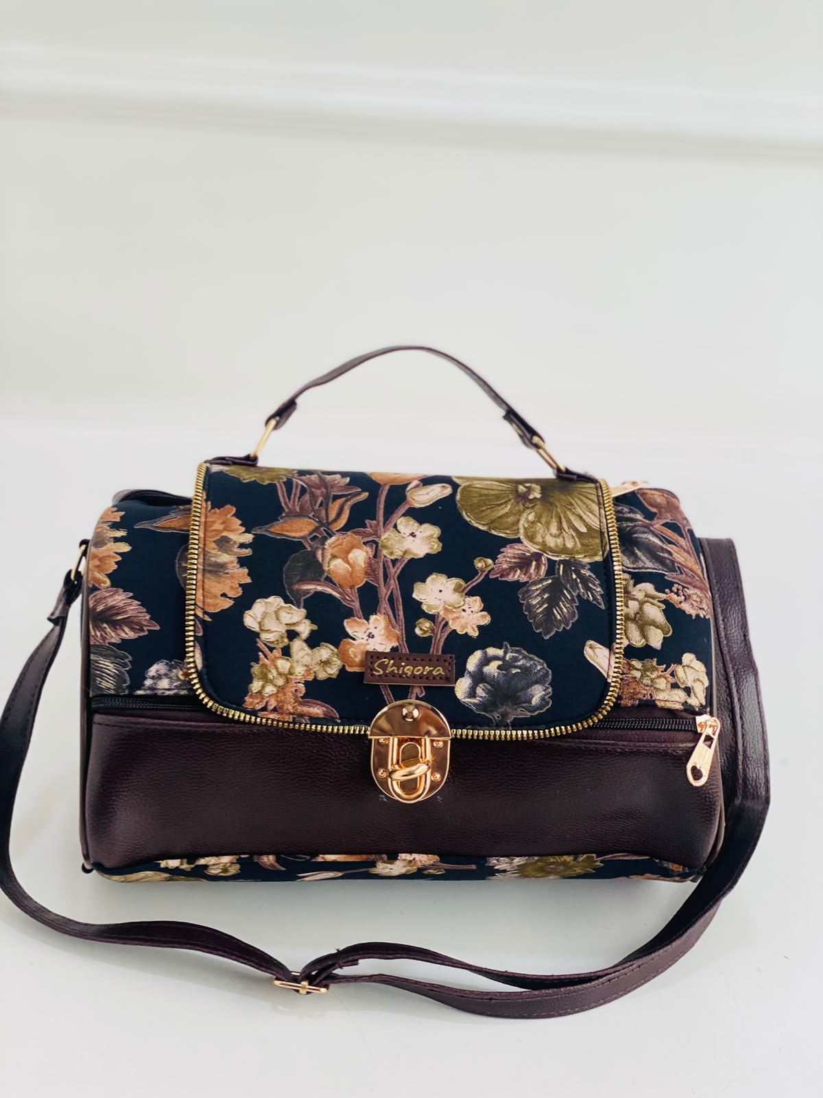 A structured handbag with a dark floral patterned flap and base, combined with a solid dark brown leather lower section and handle. It features gold-tone hardware, including a central turn-lock clasp and zipper pulls, and is displayed on a clean white background.