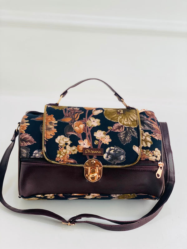 A structured handbag with a dark floral patterned flap and base, combined with a solid dark brown leather lower section and handle. It features gold-tone hardware, including a central turn-lock clasp and zipper pulls, and is displayed on a clean white background.